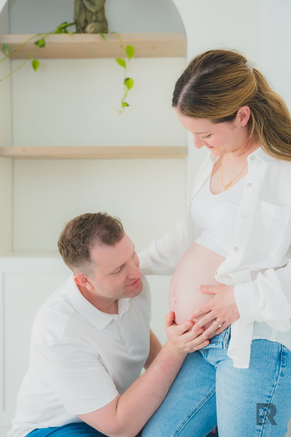 At-home maternity photoshoot pose — partner kneeling with hands on the bump